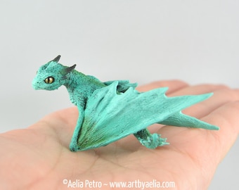 Op de afbeelding: Een teal-gekleurde drakenfiguur met gedetailleerde vleugels en een getextureerd lichaam. De draak heeft gele ogen en zwarte hoorns. De figuur wordt op een hand getoond, wat de grootte en het ingewikkelde ontwerp laat zien. Fantasy-thema verzamelobject.