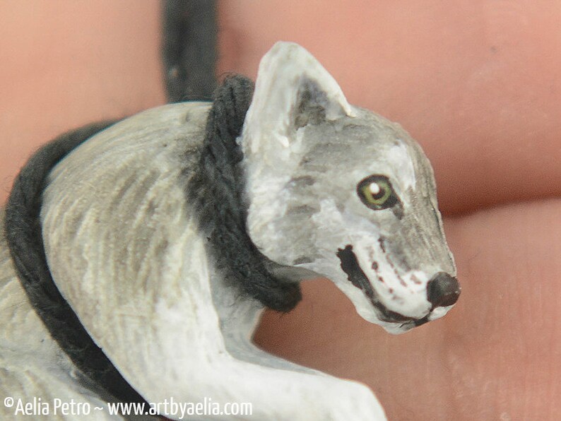 May include: A hand-painted wooden wolf figurine with a gray and white coat, black eyes, and a black rope around its neck.