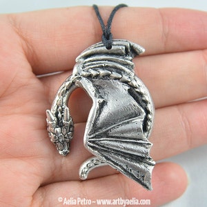 May include: Silver pendant necklace featuring a detailed dragon design with wings outstretched. The dragon is curled in a circle with its head facing forward. The necklace is on a black cord.