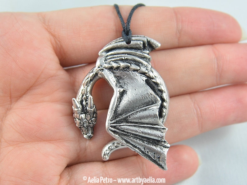 May include: Silver pendant necklace featuring a detailed dragon design with wings outstretched. The dragon is curled in a circle with its head facing forward. The necklace is on a black cord.