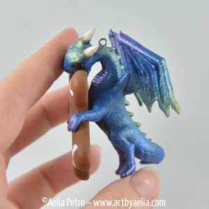 May include: A blue dragon figurine with iridescent wings and accents. The dragon is holding a brown object with white details. The figurine has a metallic finish and is a fantasy-themed item.