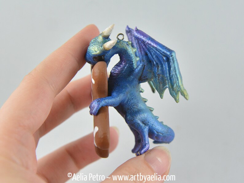 May include: A blue dragon figurine with iridescent wings and accents. The dragon is holding a brown object with white details. The figurine has a metallic finish and is a fantasy-themed item.