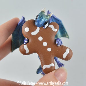 May include: A whimsical figurine of a blue and green dragon holding a gingerbread man. The dragon has iridescent wings and is holding the gingerbread man with its tiny purple claws. The gingerbread man is brown with white icing.