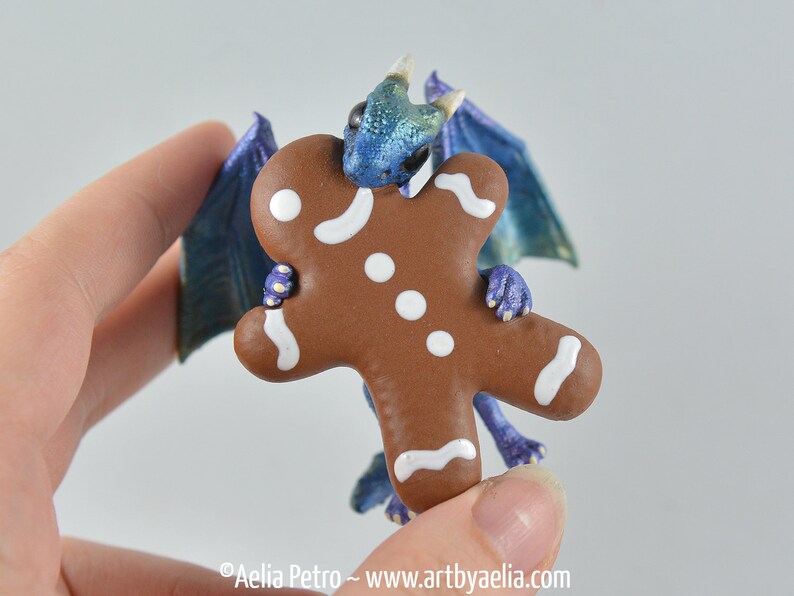 May include: A whimsical figurine of a blue and green dragon holding a gingerbread man. The dragon has iridescent wings and is holding the gingerbread man with its tiny purple claws. The gingerbread man is brown with white icing.