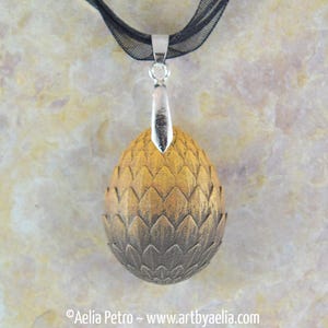 May include: A pendant necklace featuring a detailed, egg-shaped charm with a textured, scale-like design. The charm transitions from a golden hue at the top to a darker, bronze color at the bottom. It hangs from a black ribbon.