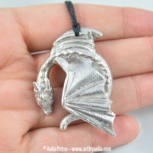 May include: Silver dragon pendant necklace with black cord. The dragon is curled up with its wings spread out.