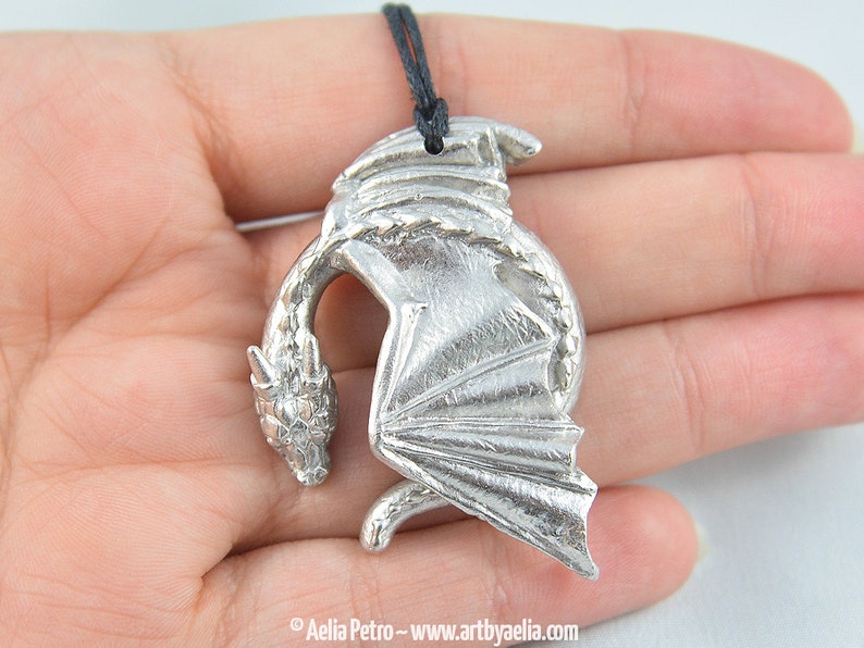 May include: Silver dragon pendant necklace with black cord. The dragon is curled up with its wings spread out.