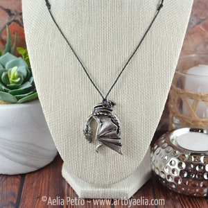 May include: A silver dragon pendant necklace on a black cord. The dragon is curled up with its wings spread out. The necklace is displayed on a white mannequin.