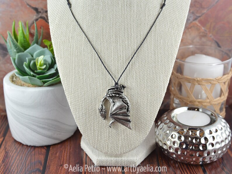 May include: A silver dragon pendant necklace on a black cord. The dragon is curled up with its wings spread out. The necklace is displayed on a white mannequin.