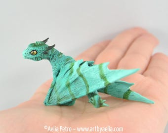 Op de afbeelding: Een kleine, zeegroene drakenfiguur met gedetailleerde vleugels en een getextureerd lichaam. De draak heeft een groen en geel oog en wordt in een hand gehouden. De figuur is een fantasy-thema verzamelobject.