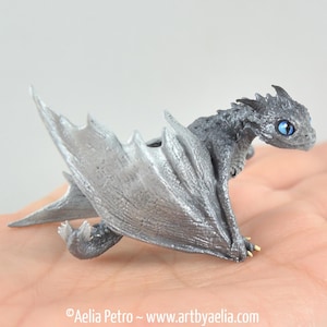 May include: A detailed figurine of a small, silver dragon with large, textured wings and bright blue eyes. The dragon is posed with its wings spread, showcasing intricate details. The sculpture is a fantasy-themed collectible.