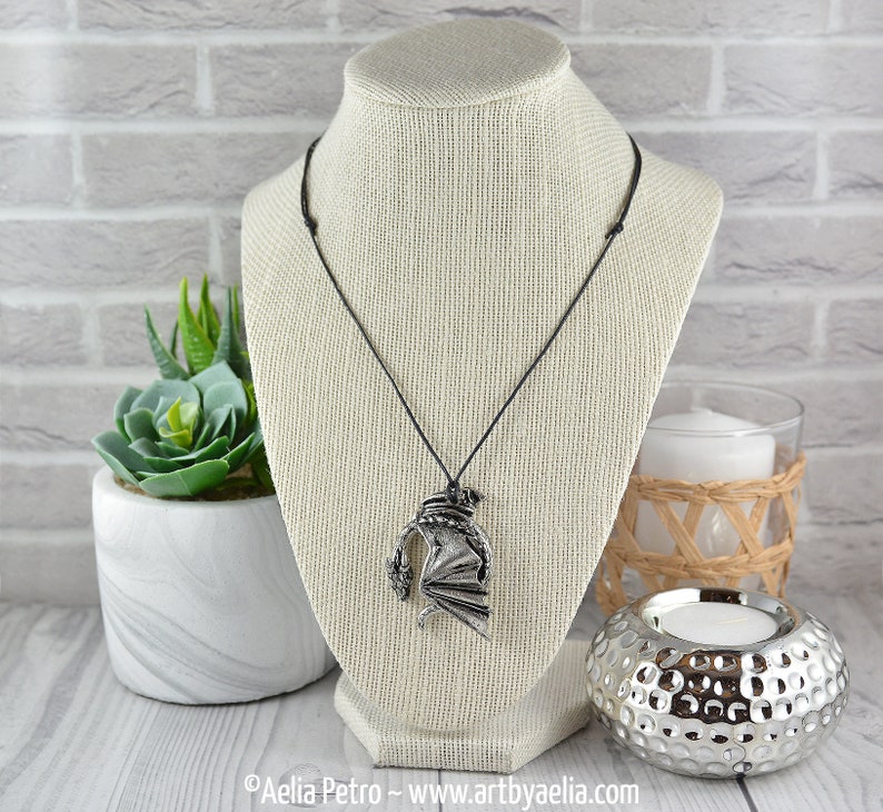 May include: A silver dragon pendant necklace on a black cord. The dragon is in a curled position with its wings spread out. The necklace is displayed on a white mannequin.