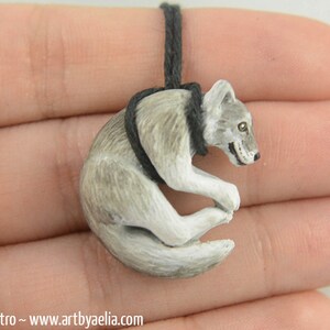 May include: A small, gray and white wolf pendant, curled up in a sleeping position, hangs from a black cord.