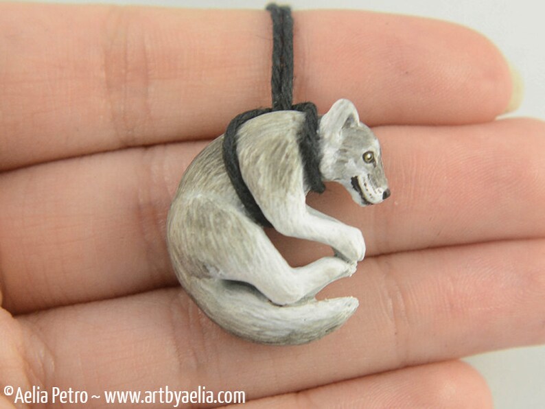 May include: A small, gray and white wolf pendant, curled up in a sleeping position, hangs from a black cord.