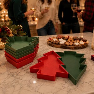 May include: A stack of green Christmas tree-shaped plates and two individual plates in red and green. The plates have raised edges and a flat base. These festive serving dishes are arranged on a light-colored countertop alongside holiday decor.