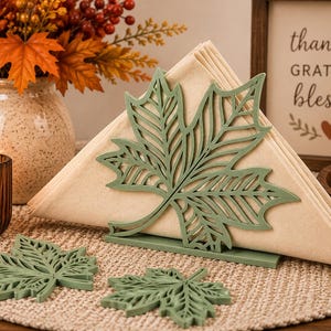 May include: A sage green metal napkin holder shaped like a maple leaf, holding folded beige napkins. Two matching sage green maple leaf coasters are placed in front of the holder on a textured surface.