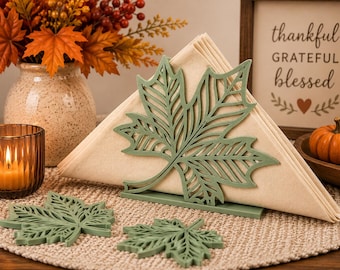 May include: A sage green metal napkin holder shaped like a maple leaf, holding folded beige napkins. Two matching sage green maple leaf coasters are placed in front of the holder on a textured surface.
