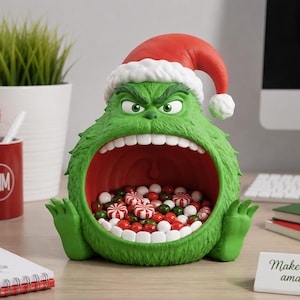 May include: A green, furry, creature-shaped bowl with a wide-open mouth filled with red, white, and green candies. The figure has a scowling expression, prominent white teeth, and wears a red and white Santa hat. The bowl is made of a textured material.