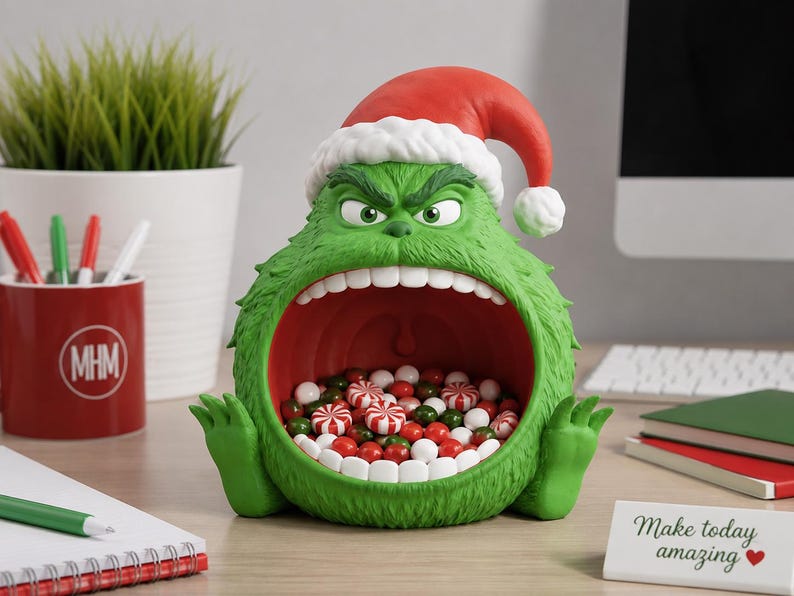 May include: A green, furry, creature-shaped bowl with a wide-open mouth filled with red, white, and green candies. The figure has a scowling expression, prominent white teeth, and wears a red and white Santa hat. The bowl is made of a textured material.