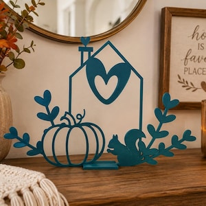 May include: A teal metal cutout decoration featuring a house outline with a heart-shaped cutout, a pumpkin, a squirrel, and leafy branches. The house has a small heart on the chimney. The piece stands on a rectangular base.