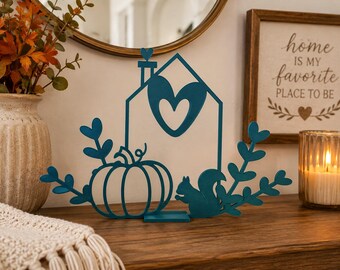 May include: A teal metal cutout decoration featuring a house outline with a heart-shaped cutout, a pumpkin, a squirrel, and leafy branches. The house has a small heart on the chimney. The piece stands on a rectangular base.