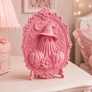 May include: A pink oval-shaped decorative plaque featuring a raised relief of a ghost wearing a witch hat, flanked by two pumpkins and floral accents. The frame is ornate with scrollwork, a crescent moon, and stars at the top.