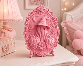 May include: A pink oval-shaped decorative plaque featuring a raised relief of a ghost wearing a witch hat, flanked by two pumpkins and floral accents. The frame is ornate with scrollwork, a crescent moon, and stars at the top.