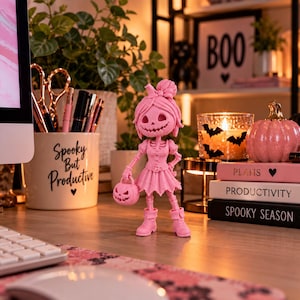 May include: A pink skeleton figurine with a bow in its hair, wearing a dress and boots, holding a small pumpkin bucket. The figurine stands on a desk near a mug labeled Spooky But Productive and a stack of books labeled Plans, Productivity, and Spooky Season.