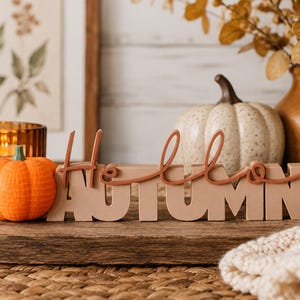 May include: A decorative sign featuring the word AUTUMN in block letters with the word Hello in a cursive script layered on top. The sign is beige with rust-colored lettering and is displayed alongside a small orange pumpkin.