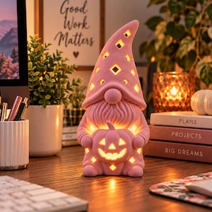 May include: A pink ceramic gnome figurine with a tall, pointed hat featuring diamond-shaped cutouts. The gnome has a textured beard and holds a carved pumpkin with a jack-o'-lantern face. The figure is illuminated from within, casting a warm glow through the cutouts in the hat and the pumpkin.