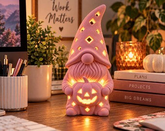 May include: A pink ceramic gnome figurine with a tall, pointed hat featuring diamond-shaped cutouts. The gnome has a textured beard and holds a carved pumpkin with a jack-o'-lantern face. The figure is illuminated from within, casting a warm glow through the cutouts in the hat and the pumpkin.