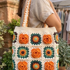 May include: A crochet tote bag made of off-white yarn featuring a pattern of square motifs. The design alternates between flat floral squares in shades of dark green, brown, and light green, and squares with three-dimensional orange roses.