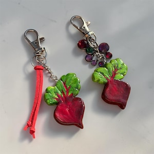 May include: Two keychains featuring beet charms with deep red roots and vibrant green leaves. One keychain includes a silver lobster clasp, a short red suede cord, and a silver chain. The other features a silver lobster clasp and a cluster of small, round, multicolored faceted beads.