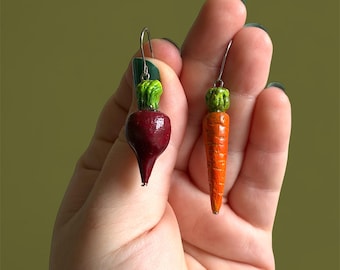 May include: A pair of dangle earrings featuring miniature vegetable charms. One earring is a deep red beet with green leafy tops, and the other is a bright orange carrot with green leafy tops. Both charms hang from silver-toned metal hooks.