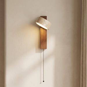 Könnte beinhalten: Eine Wandleuchte mit einer vertikalen, rechteckigen Rückplatte aus dunklem Holz und einem geneigten, zylindrischen Lampenschirm mit strukturierter, beigefarbener Steinoberfläche. Die Lampe strahlt warmes Licht aus und hat eine Zugkette.
