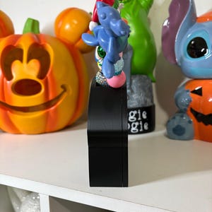 Peut inclure: Une base rectangulaire noire surmontée d'un arrangement vertical de perles colorées, incluant une figurine de personnage bleu portant un chapeau rouge. Les perles présentent diverses textures, dont des finitions lisses, pailletées et métalliques.