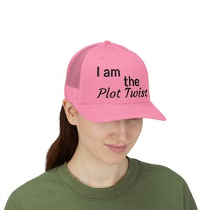 May include: A bright pink trucker cap featuring a mesh back and a solid front panel. The text "I am the Plot Twist" is printed in black lettering across the front of the cap, with "I am" and "the" in a sans-serif font and "Plot Twist" in script.