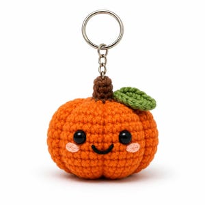 May include: A crocheted orange pumpkin keyring featuring a brown stem, a single green leaf, two black plastic eyes, a stitched black smile, and two small pink blush marks on the cheeks, attached to a silver metal split ring and chain.