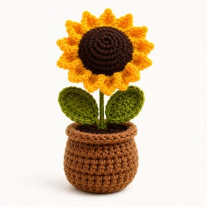 Può includere: Un girasole all'uncinetto in un vaso marrone. Il fiore ha un centro marrone scuro con petali giallo brillante. Due foglie verdi sono attaccate al gambo centrale. Il vaso è realizzato in filato marrone con un motivo a maglia a coste.