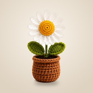 Puede incluir: Una flor de margarita tejida a ganchillo con pétalos blancos y un centro amarillo brillante, con dos hojas verdes en un tallo central. La flor está plantada en una maceta de ganchillo marrón texturizada con un borde grueso y base sólida.