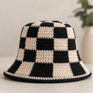 May include: A crochet bucket hat featuring a repeating checkerboard pattern in alternating black and off-white yarn. The hat has a rounded crown and a downward-sloping brim, constructed with a visible textured stitch throughout the entire piece.