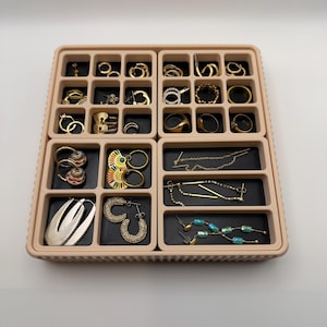 May include: A beige plastic jewelry organizer tray with multiple square and rectangular compartments containing various gold-toned earrings, rings, and dangling jewelry pieces. The tray features a black velvet-like lining in each section.
