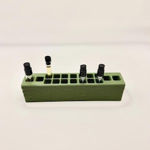 May include: A rectangular olive green storage stand with a textured surface and multiple square slots. Four small, clear glass vials with black caps are inserted into the slots, while the remaining openings in the stand are empty.
