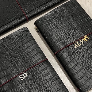 Peut inclure: Trois carnets rectangulaires noirs avec un motif texturé en peau de crocodile. Chaque carnet est maintenu par un fin élastique rouge foncé. Deux carnets présentent des breloques métalliques sur l'élastique : l'une avec les lettres SD et l'autre ALYX.