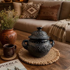 Puede incluir: Una olla de cerámica negra con tapa, dos asas laterales y patrones florales y de puntos en relieve. La olla se asienta sobre un mantel individual redondo de yute trenzado con flecos. La superficie es de color negro mate con detalles decorativos.
