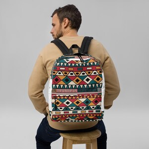 May include: A backpack featuring a repeating geometric pattern in red, teal, yellow, and white. The design includes rows of triangles, diamonds, and horizontal lines. The bag has black shoulder straps and a black zipper pull on the front pocket.