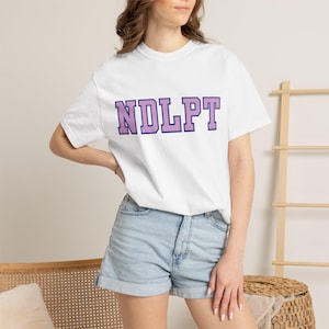 May include: A white short-sleeved t-shirt featuring the text NDLPT in large, block-style lavender letters with a dark purple outline across the chest. The shirt is paired with light blue denim shorts that have a cuffed hem at the bottom.