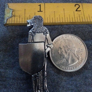 Puede incluir: Un pin de metal plateado que representa una criatura parecida a un lobo que lleva armadura y sostiene un escudo. El pin es aproximadamente del tama&ntilde;o de una moneda de 25 centavos.