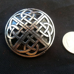 May include: A silver Celtic knot design brooch with a black background. The knot is intricate and detailed, and the brooch is a beautiful piece of jewelry.