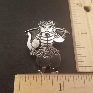 May include: A silver-colored cat warrior pin, approximately 1 inch tall, with a detailed design. The cat is depicted in samurai armor, holding a sword. The pin has a shiny, metallic finish and is set against a dark background.